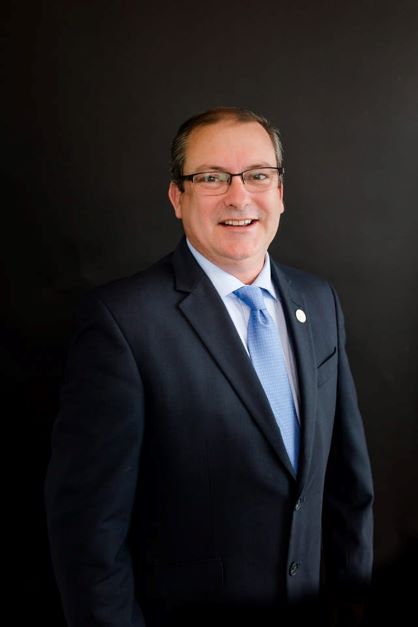 Man with dark hair wearing a black suit, with a light blue button down shirt and blue tie, standing in front of a black background.