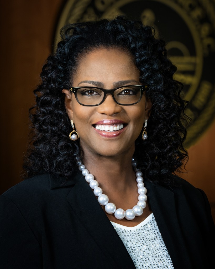 Woman with black hair and glasses, wearing a black suit jacket, white shirt and a pearl necklace.