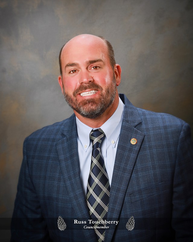 Man with brown hair and a beard. He is wearing a navy suit and tie, with a light blue button up shirt, and smiling at the camera. 