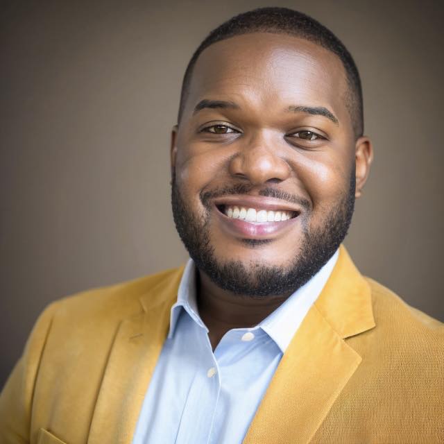 Man wearing a yellow suit jacket with a light blue shirt, smiling at the camera.